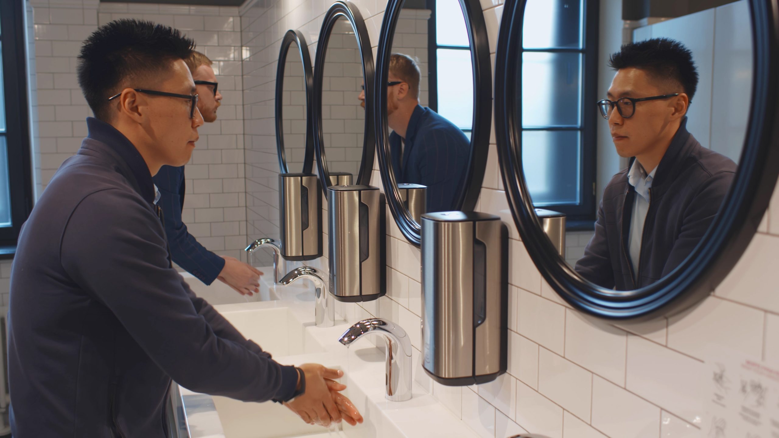 Office building bathroom design benefits all patrons, such as this young adult man washing his hands in front of a commercial restroom mirror.